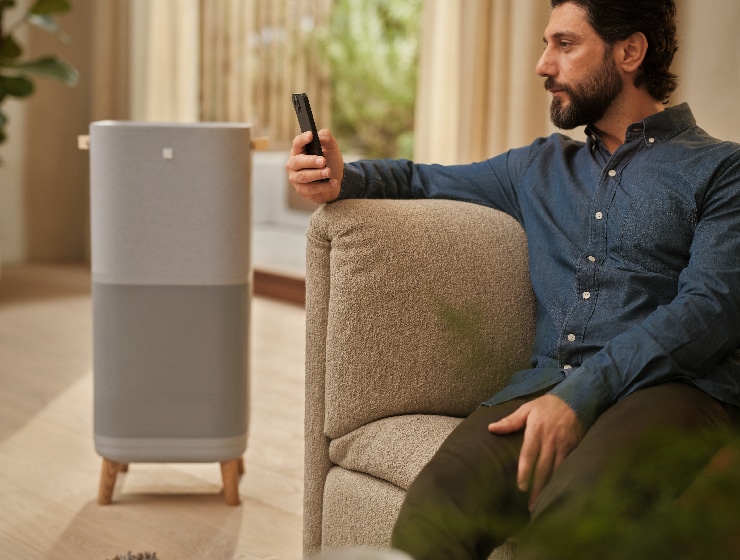 Vīrietis ar telefonu sēž blakus gaisa attīrītājam.