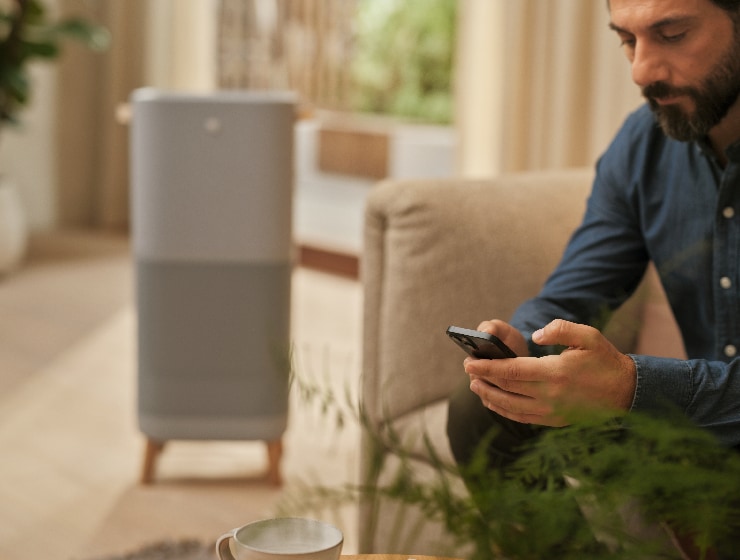 Vīrietis ar telefonu sēž blakus gaisa attīrītājam.