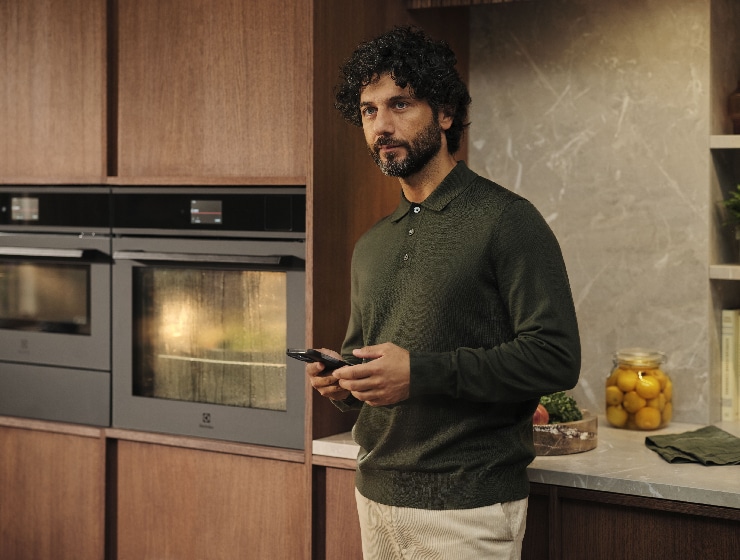 Vīrietis stāv virtuvē un tur rokā telefonu.