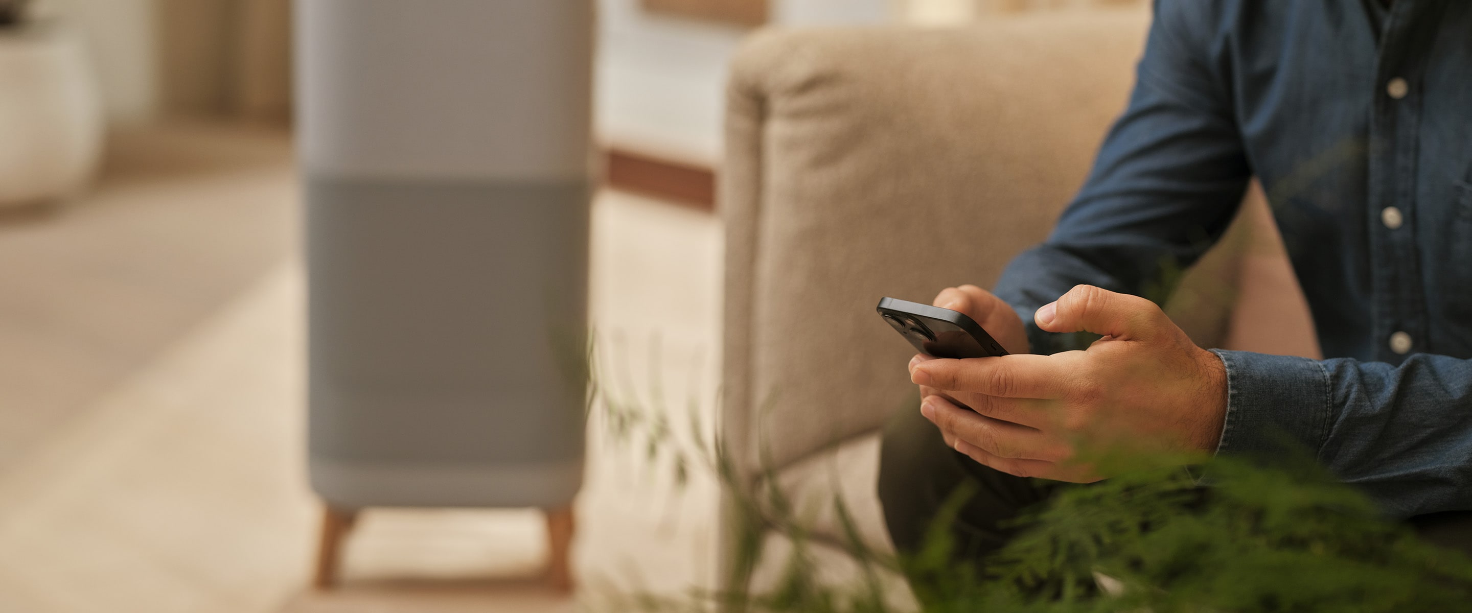 Vīrietis sēž un tur telefonu rokā. Fonā gaisa attīrītājs.